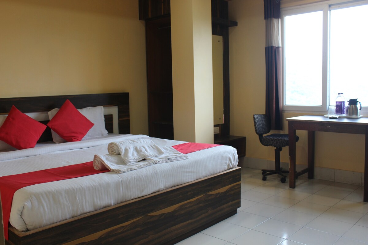 A spacious room is centered around a large bed adorned with crisp white linens and red accent pillows. A small desk and chair are situated next to a window that allows natural light to fill the space, while neatly folded towels are placed on the bed.