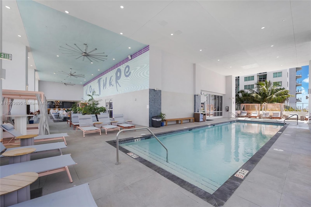 A modern pool area is highlighted with several lounge chairs set around a rectangular swimming pool. The space features large windows and tropical plants, creating a bright atmosphere. Ceiling fans are visible above, adding a touch of comfort to this inviting communal area.