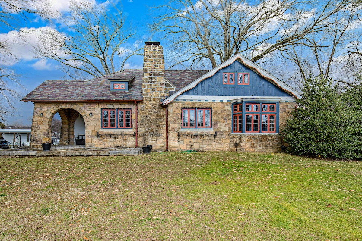 The exterior of The Howe House is presented, showcasing a historic stone façade with large, multi-paned windows. An arched entryway adds character, while a well-maintained lawn extends in front. Trees provide a natural backdrop, enhancing the serene outdoor setting.
