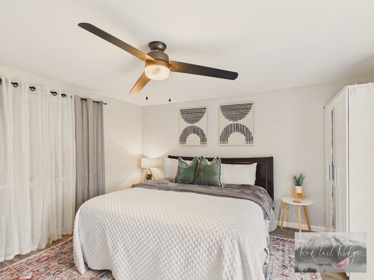 A spacious bedroom is furnished with a large bed covered in a textured blanket. Decorative wall art adorns the light-colored walls, while sheer curtains allow natural light to filter in. A modern ceiling fan and bedside lamp enhance the comfort of the room.