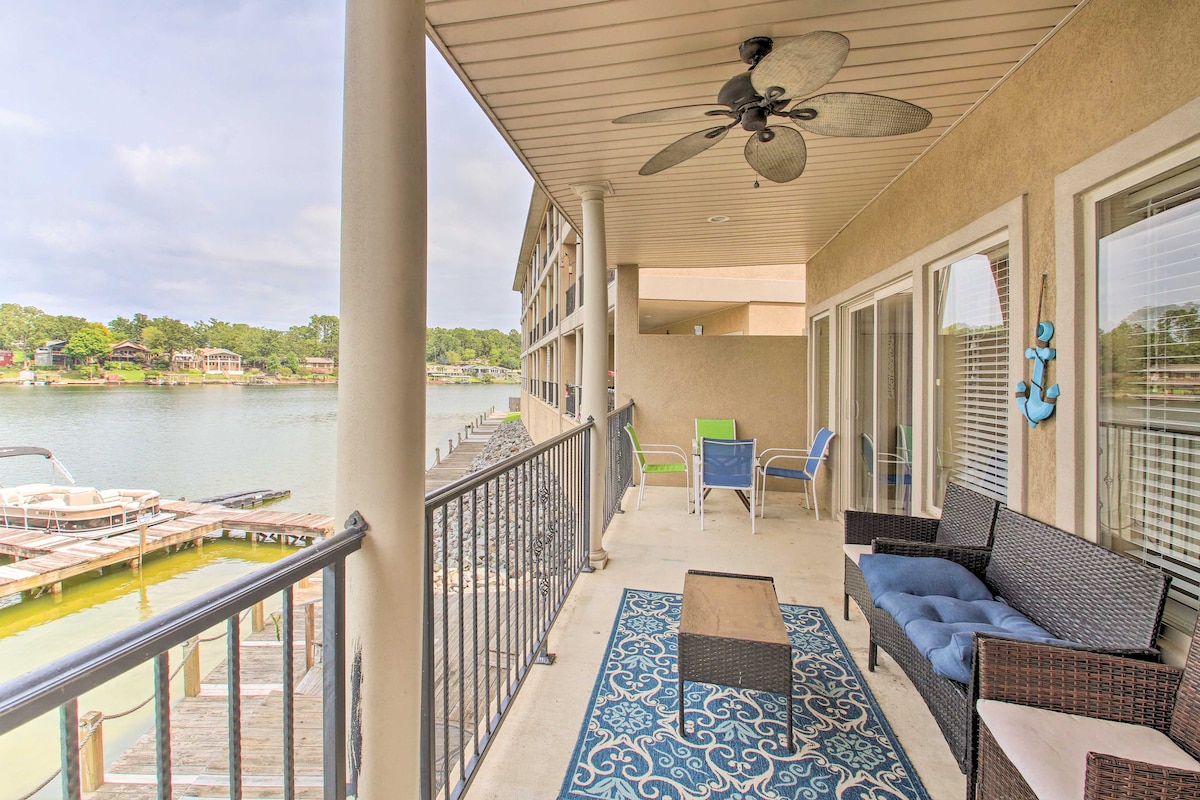 A spacious covered balcony features outdoor seating, including a wicker couch and colorful chairs. A light rug rests on the floor, while a ceiling fan hangs above. Views of Lake Hamilton and nearby boat slips are visible beyond the railing.