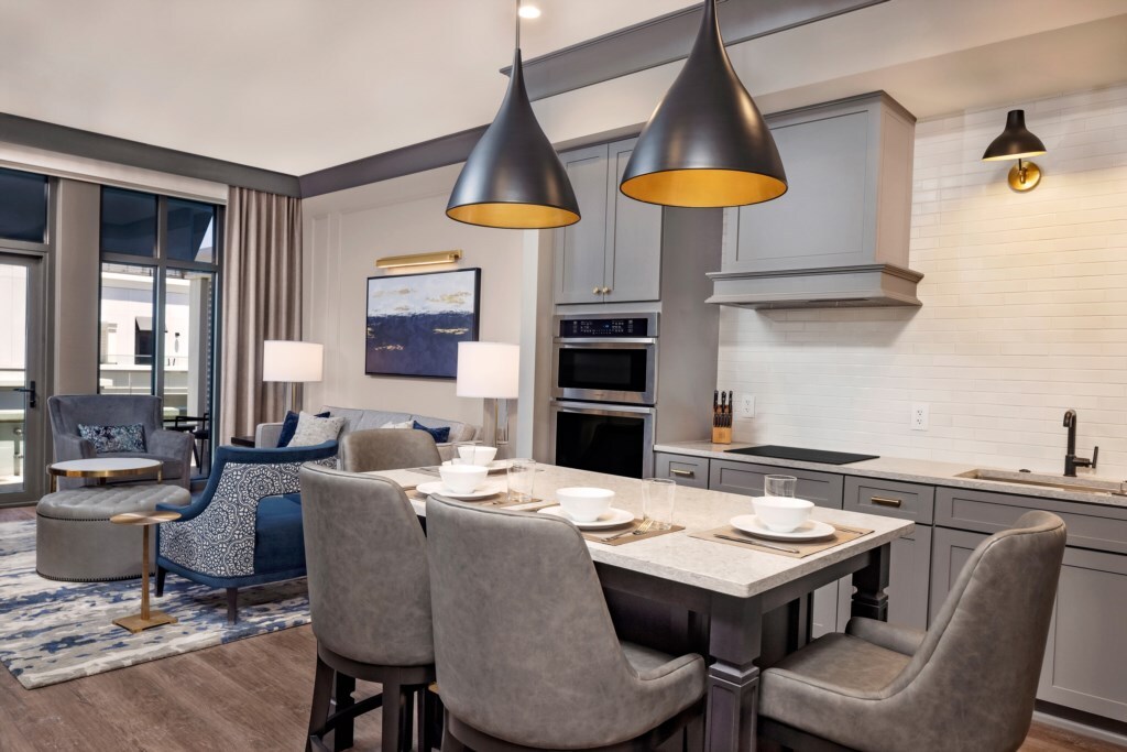 A modern kitchen and dining area are highlighted, featuring elegant pendant lights above a spacious table set for four. Gray cabinets complement marble countertops, while a comfortable living area is visible in the background, showcasing stylish seating and large windows providing natural light.