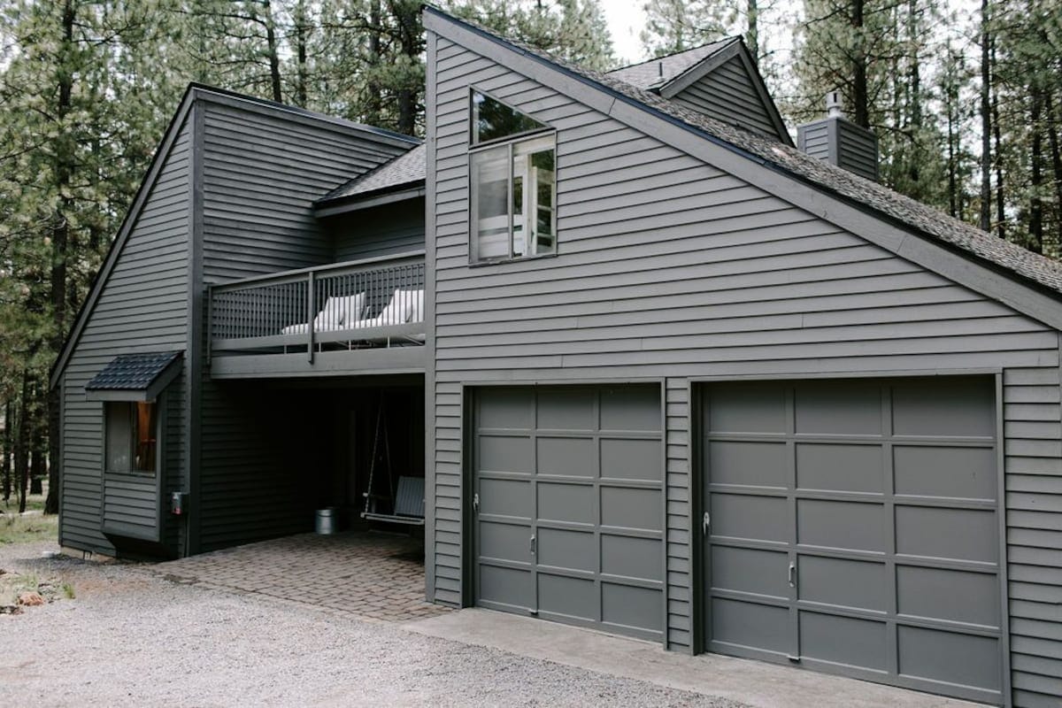 A modern two-story cabin is set among towering pine trees. The exterior features grey siding and a balcony with two white lounge chairs. A spacious driveway leads to two garage doors, complemented by a stone pathway at the front.