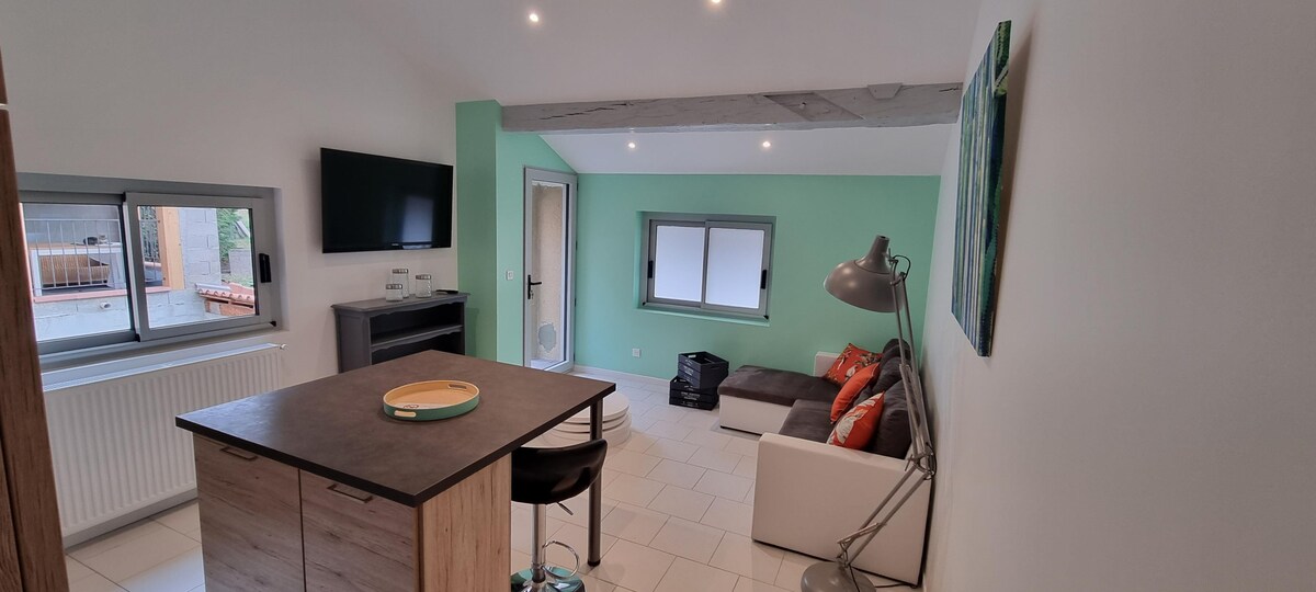 A modern living area features a light-colored sofa with colorful cushions. A flat-screen TV is mounted on one wall, and a compact dining area is set nearby. Natural light enters through large windows, while a door leads to an outdoor space.