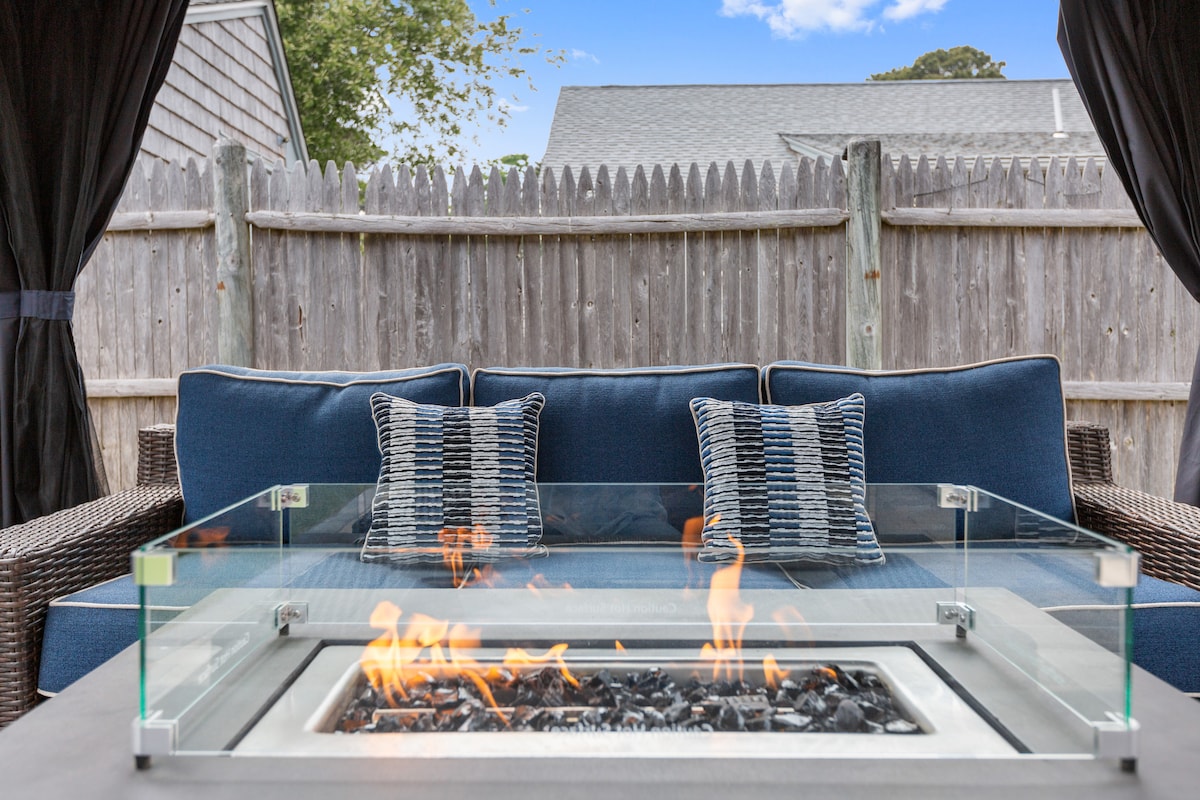 An outdoor seating area features a blue cushioned sofa with decorative throw pillows, positioned in front of a glass-top propane fire pit. The surrounding fenced backyard offers privacy with a wooden fence, enhancing the cozy outdoor atmosphere.