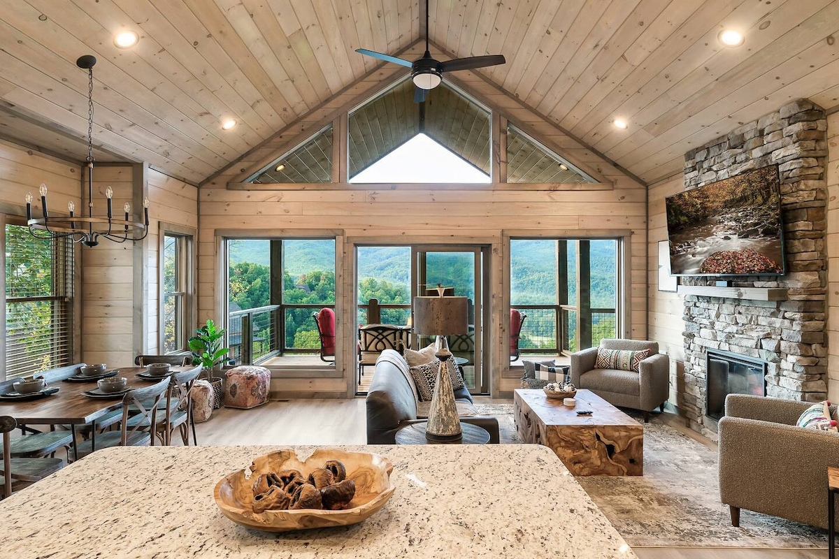 The open-concept living space features high vaulted ceilings and large windows that showcase expansive mountain views. A cozy seating area is arranged around a stone fireplace, while a dining table and kitchen island invite communal dining. Natural wood tones create a calming ambiance.