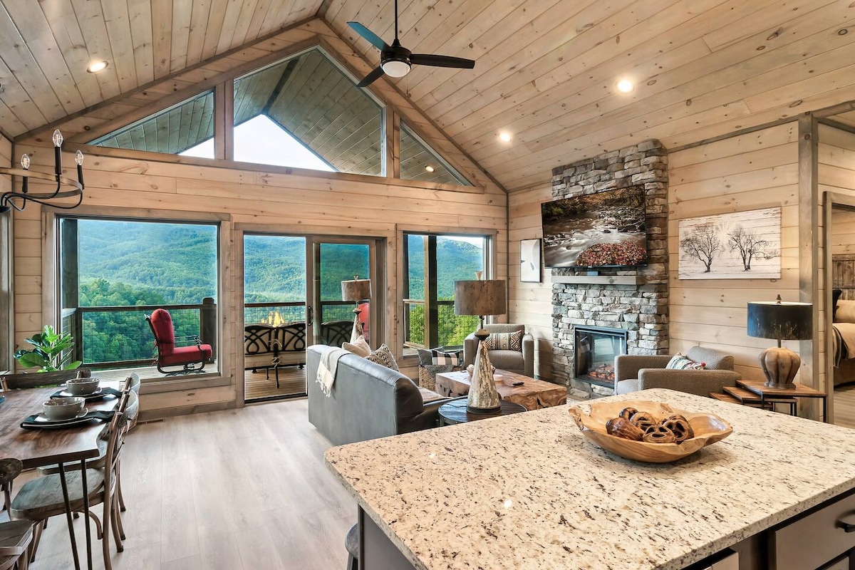 An open living space features high vaulted ceilings and a stone fireplace, with large windows providing panoramic mountain views. A dining area with a wooden table is visible, alongside a kitchen island topped with granite. Neutral-toned furnishings complement the inviting atmosphere.