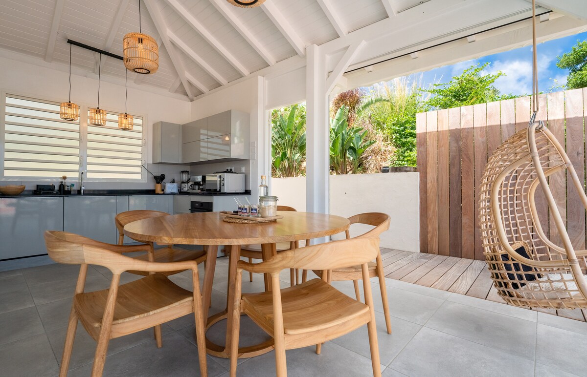 A dining area features a round wooden table surrounded by four lightweight chairs. The space is designed with an open concept, leading to an outdoor area visible through large sliding doors. Natural light illuminates the room, highlighting the minimalist decor.