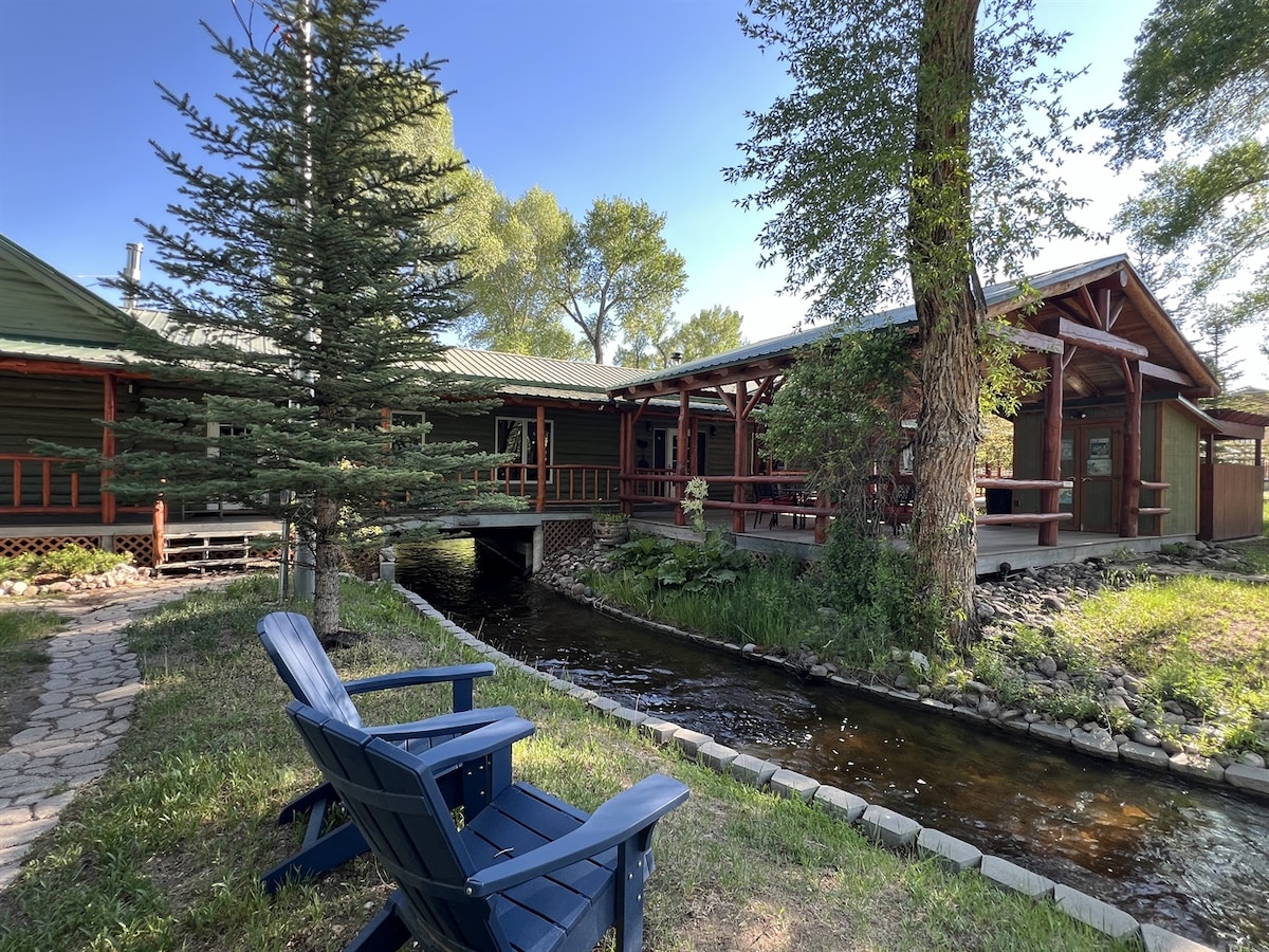 A picturesque view of the lodge showcases a winding stream lined with stones and green grass. Two blue adirondack chairs invite relaxation beside the water, while the expansive structure of the log home is visible, surrounded by trees and a clear blue sky.