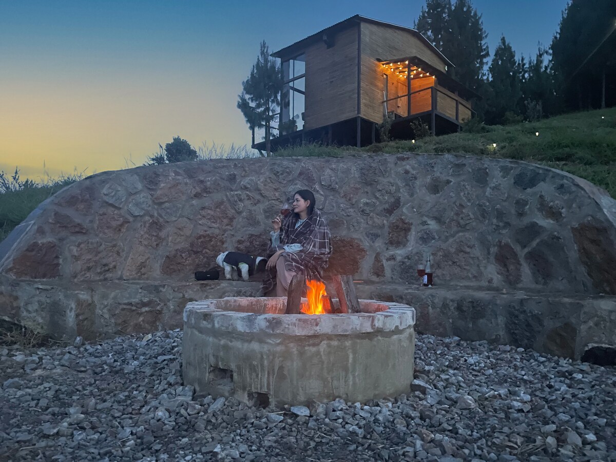 An outdoor fire pit is surrounded by stones, with warm flames creating a cozy atmosphere. A wooden cabin is visible in the background, illuminated softly by string lights. The area is decorated with natural stone and gravel, offering a serene setting for relaxation.