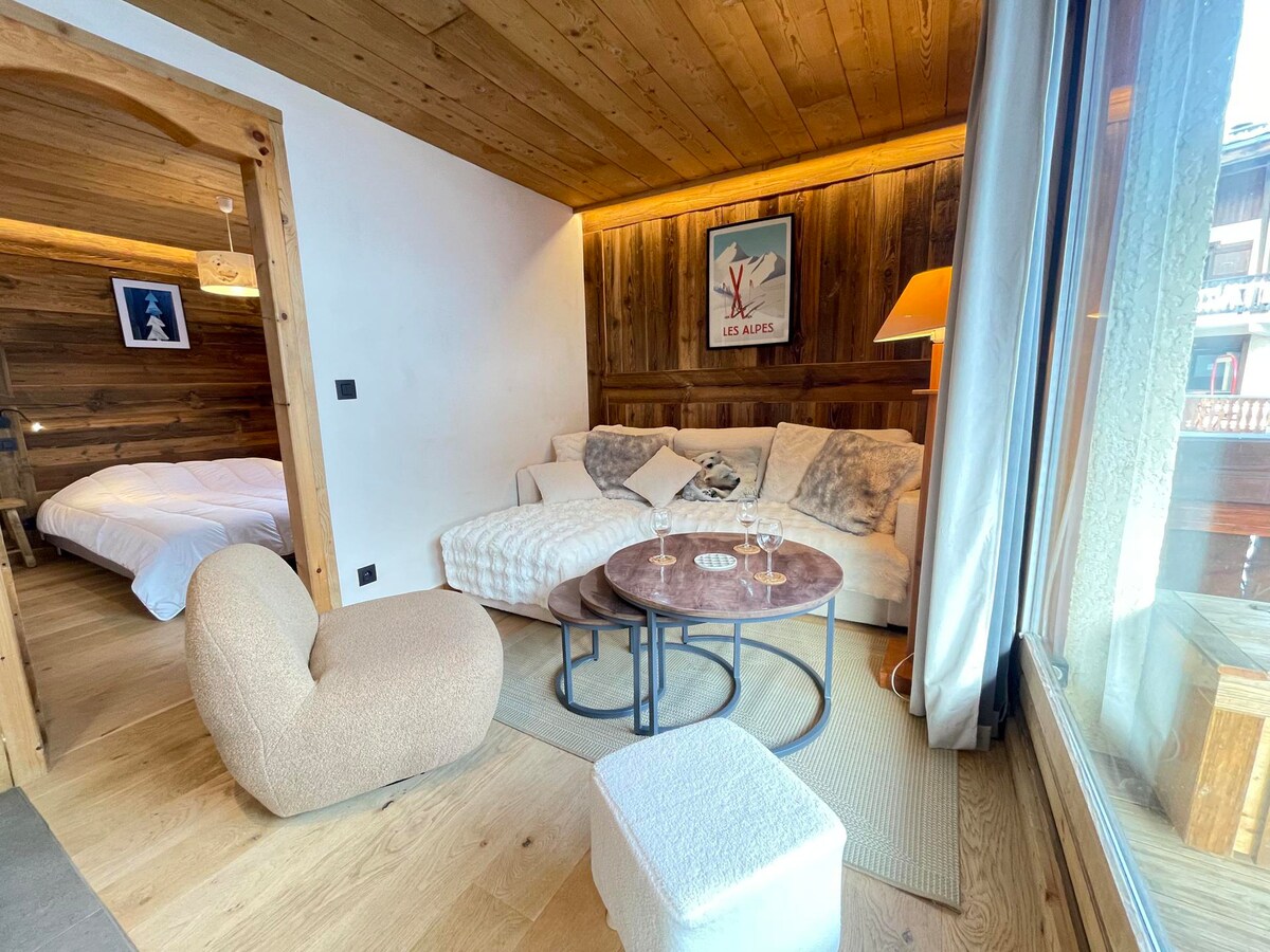 A comfortable living area features a modern sofa and two circular tables. Natural wood accent walls create a warm atmosphere. A cozy armchair invites relaxation, while large windows allow soft, natural light to illuminate the space. A glimpse of the bedroom is visible in the background.