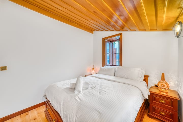 ห้องนอนเรียบง่ายและอบอุ่นพร้อมเตียงนอนสบายๆ ผ้าปูที่นอนนุ่ม และการตกแต่งด้วยไม้อันอบอุ่นทั่วทั้งห้อง หน้าต่างบานเล็กให้แสงธรรมชาติและวิวทะเลเล็กน้อย สร้างพื้นที่ที่เงียบสงบและผ่อนคลาย