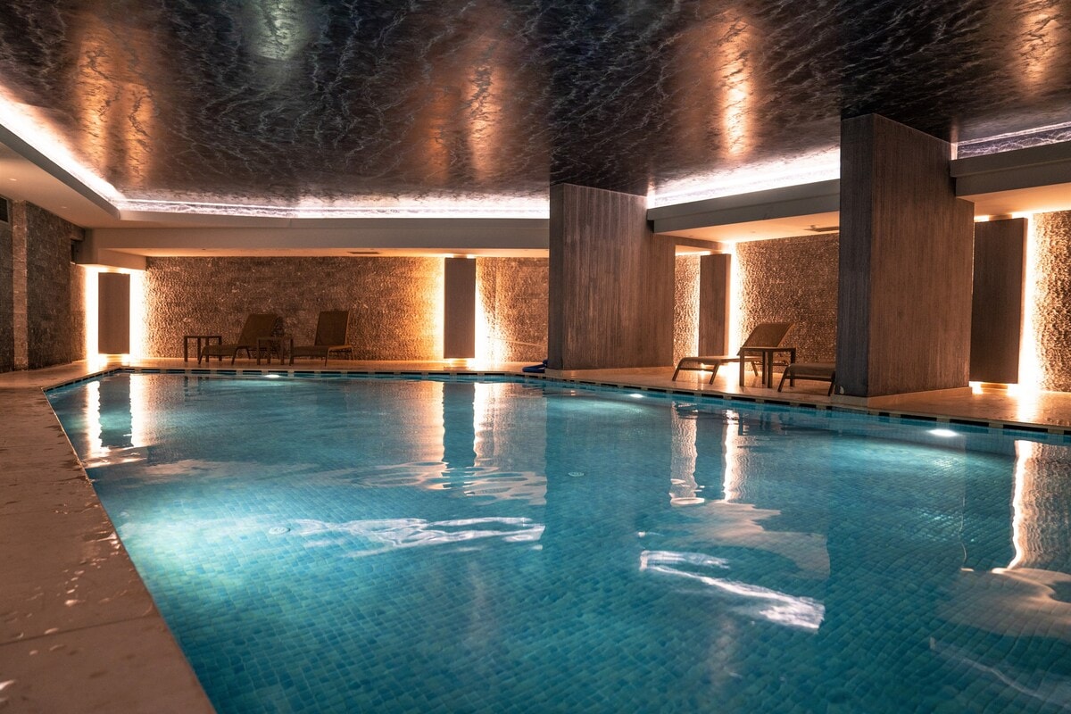 An indoor heated pool is featured, with clear blue water reflecting the softly lit ceiling. Lounge chairs are positioned around the pool area, and textured walls enhance the serene ambiance. Subtle lighting creates a calming environment, suitable for relaxation.