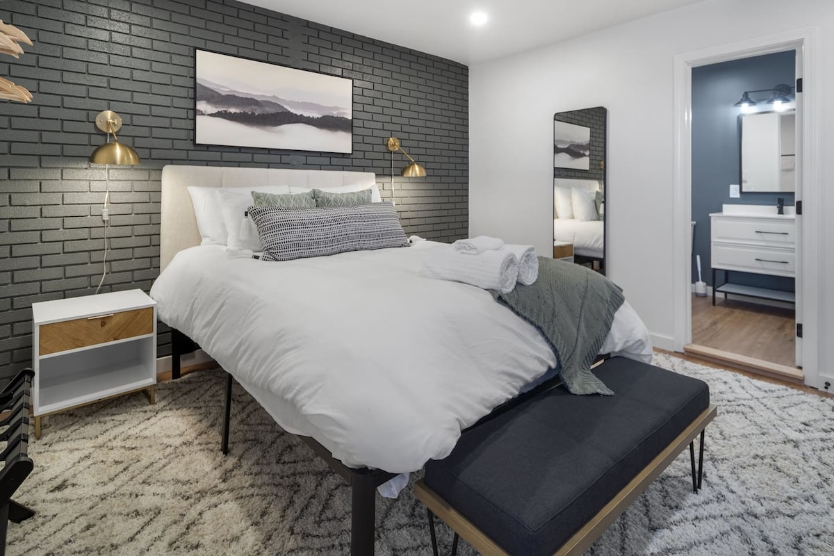 A spacious bedroom features a plush bed with crisp linens, flanked by modern bedside lamps. A sleek bench at the foot of the bed adds functionality. A large mirror reflects the room, while an adjoining bathroom is visible through an open doorway.