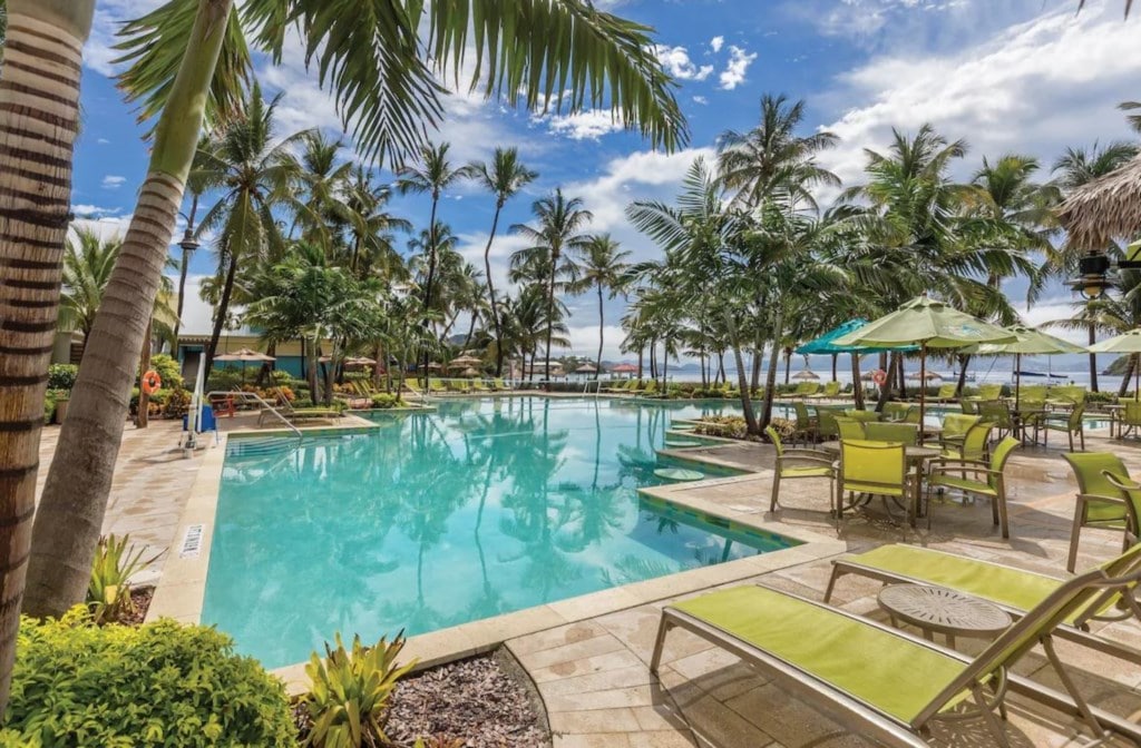 A refreshing pool is surrounded by palm trees and tropical foliage. Lounge chairs and tables with umbrellas are arranged on the deck, providing a relaxing outdoor space. The bright blue sky complements the serene environment, inviting guests to enjoy the beautiful surroundings.