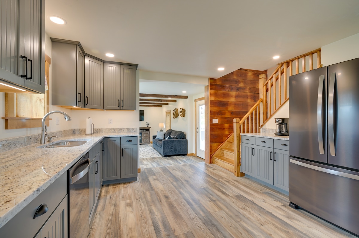 A modern kitchen features gray cabinetry and granite countertops, providing ample space for meal preparation. Stainless steel appliances are visible, including a refrigerator. The kitchen area transitions into an open living space with a staircase leading to the upper floor.