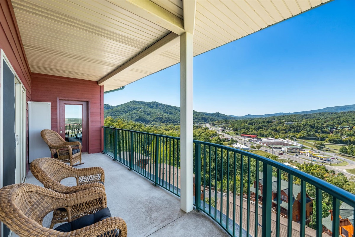 A spacious wrap-around balcony features woven chairs, offering views of the surrounding mountains and valley. The railings are designed for safety, while the scenic landscape stretches in the distance, showcasing a blend of greenery and urban features.
