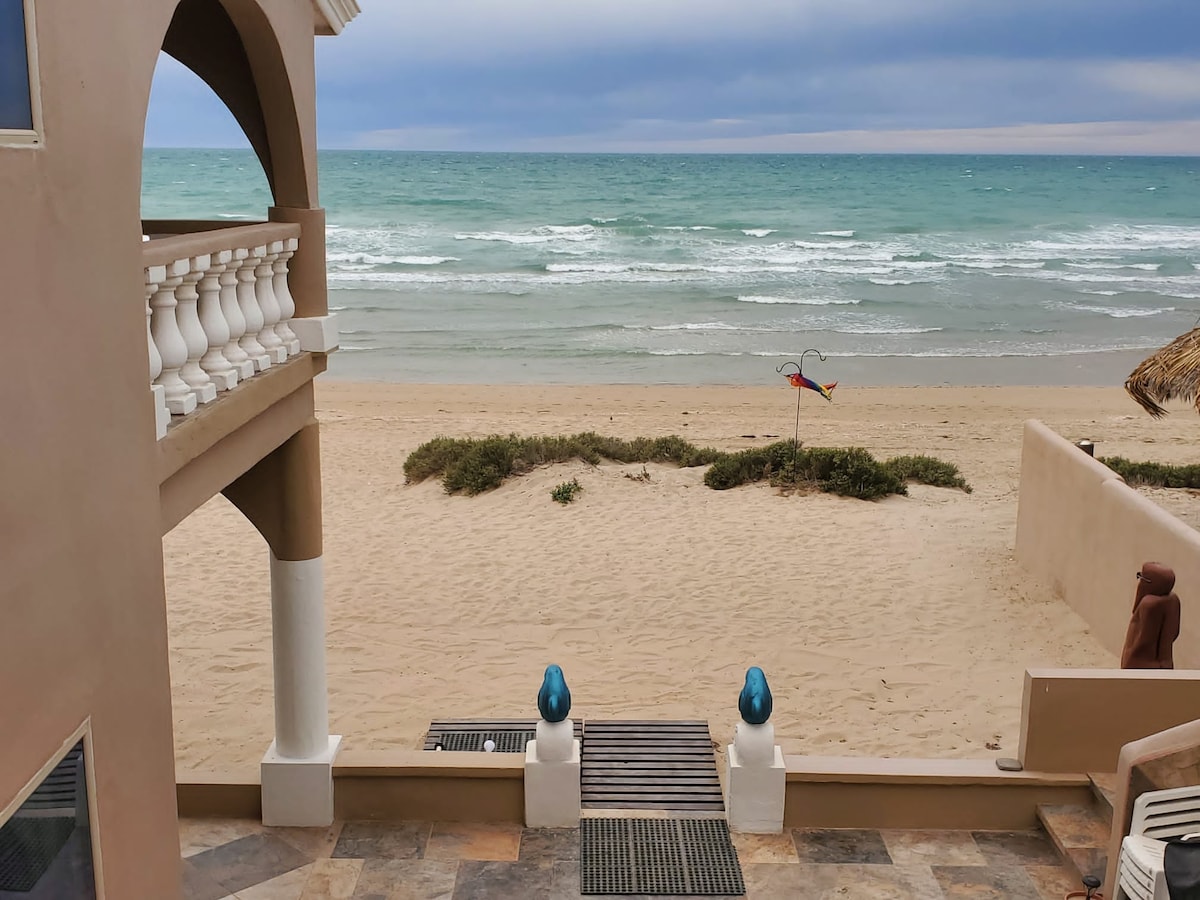 A view from the balcony reveals sandy beachfront leading to the ocean, with gentle waves lapping at the shore. The scene is complemented by light shrubbery and a colorful beach decoration in the distance. The sky displays an overcast hue, enhancing the serene coastal atmosphere.