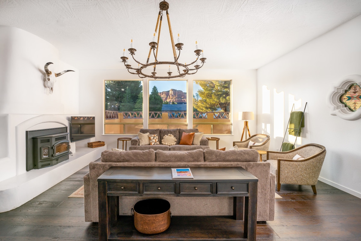 A cozy living room offers a spacious sectional sofa facing large windows that provide natural light and views of surrounding trees. A wood pellet stove sits in the corner, complemented by a wooden coffee table. A chandelier hangs above, and comfortable armchairs add to the inviting ambiance.