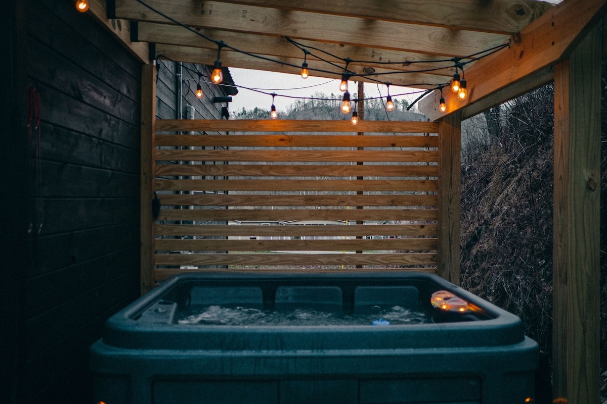 A secluded hot tub is nestled under a wooden pergola, surrounded by privacy slats. Softly glowing string lights hang overhead, creating a tranquil ambience. The inviting spa is set against a backdrop of natural greenery, enhancing the serene outdoor experience.