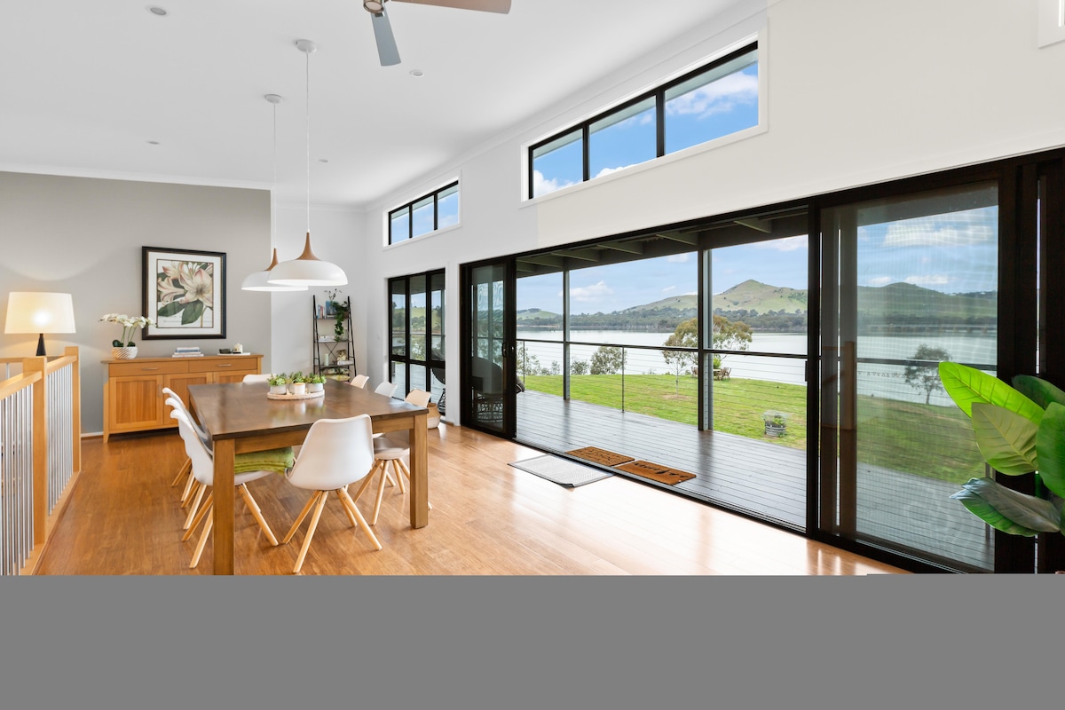 The dining area features a large table surrounded by white chairs, with a sideboard displaying decorative items. Expansive glass doors provide a view of Lake Eildon and the surrounding hills, filling the space with natural light. Green plants are seen nearby.