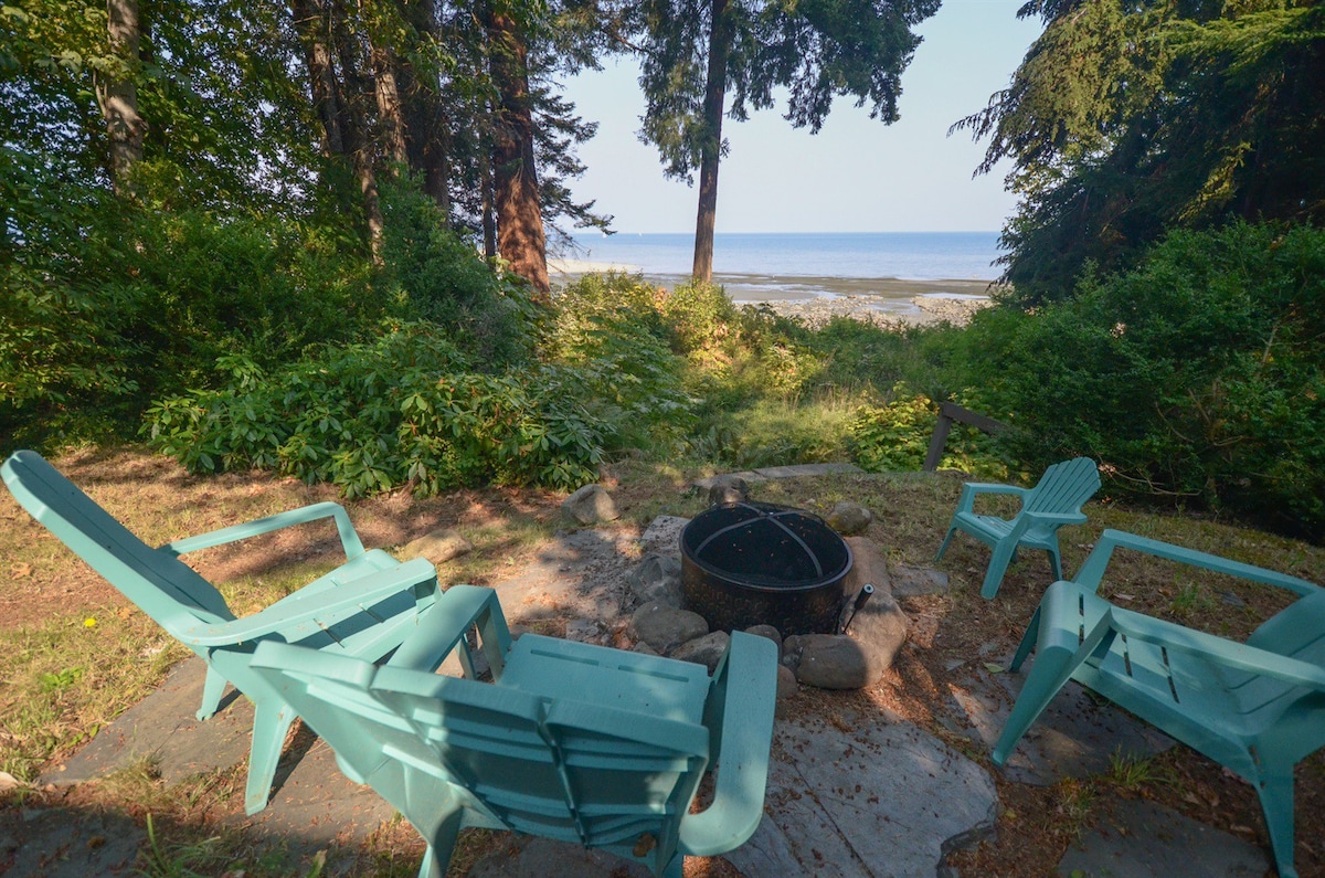 A fire pit is surrounded by several blue adirondack chairs, set on a stone patio. Lush greenery leads to a view of the ocean, with gentle waves visible in the distance. The serene setting offers a perfect spot for outdoor gatherings.