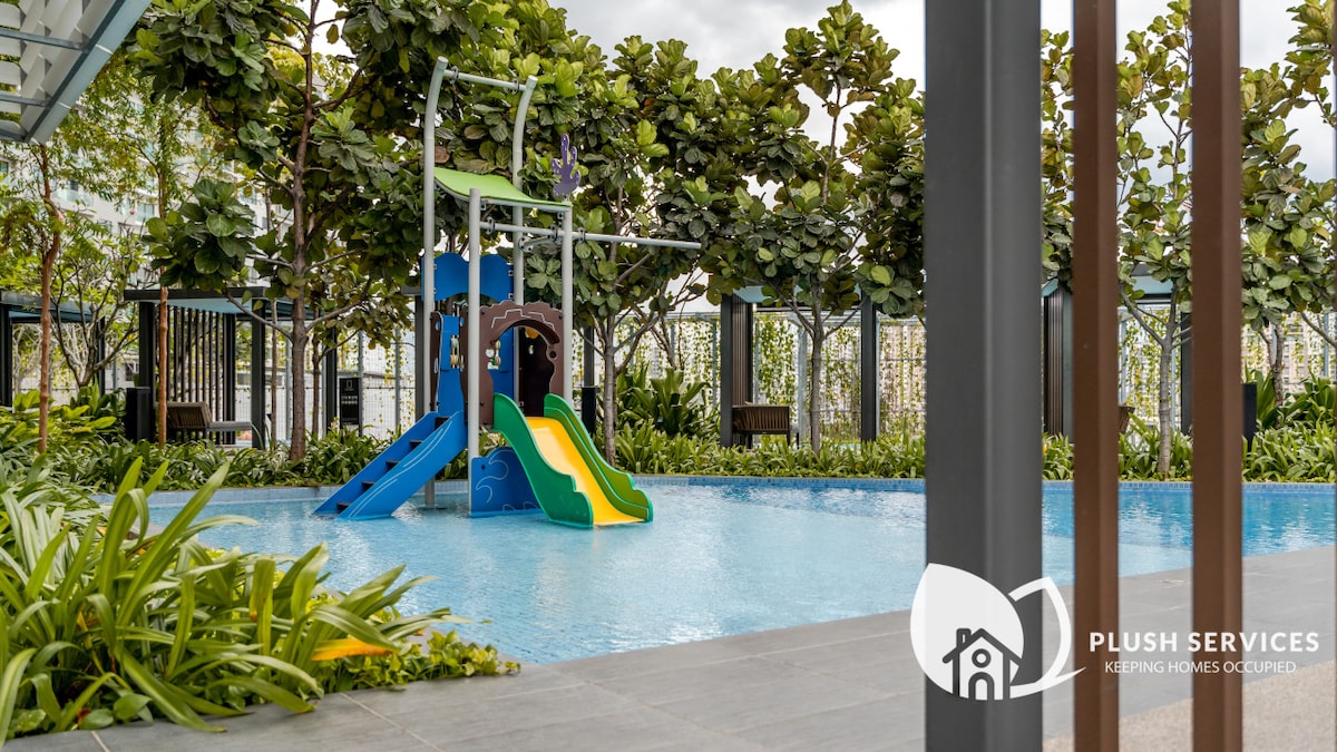 A family-friendly swimming pool area features a colorful water slide alongside a shallow children's play section. Surrounding greenery adds a lush touch to the inviting space, complemented by comfortable seating options positioned for relaxation.