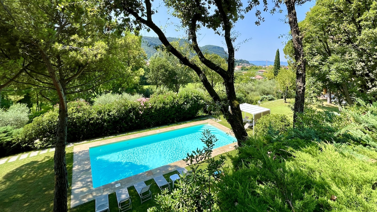 An outdoor swimming pool is surrounded by lush greenery, providing a serene escape. Comfortable lounge chairs are positioned near the poolside, inviting relaxation under the sun. The distant view includes hills and trees, blending natural beauty with a sense of privacy.