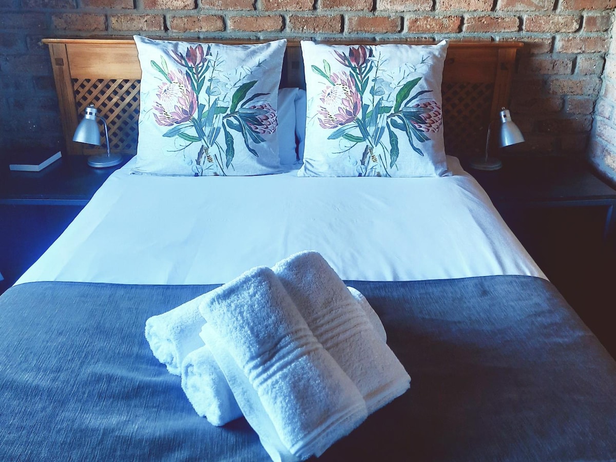 A cozy sleeping arrangement is highlighted by a spacious bed adorned with floral accent pillows. A folded set of towels is neatly placed at the foot of the bed, and subtle lighting from table lamps on either side adds to the overall comfort.