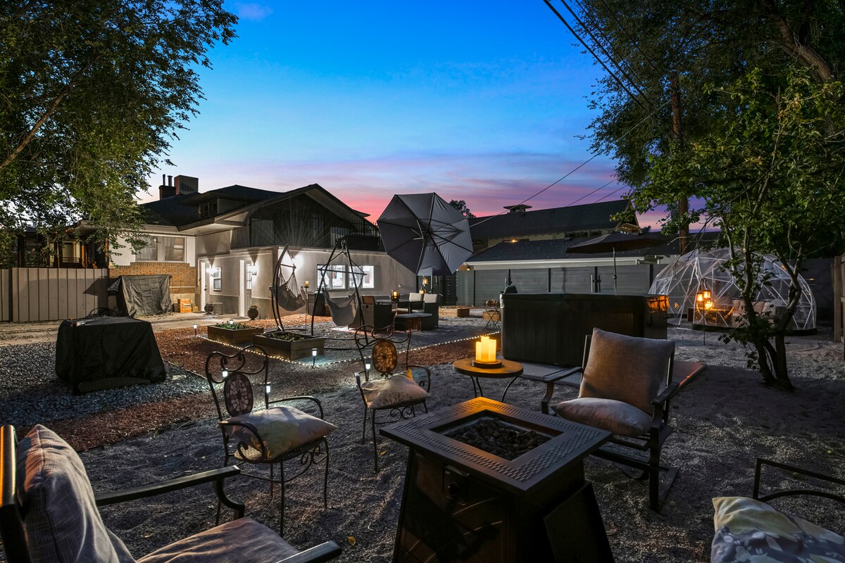 An outdoor space is illuminated by soft lighting, featuring comfortable seating around a fire pit. Several unique structures are visible in the background, along with hammocks and an inviting hot tub. The sky transitions into evening hues of blue and purple, creating a serene ambiance.