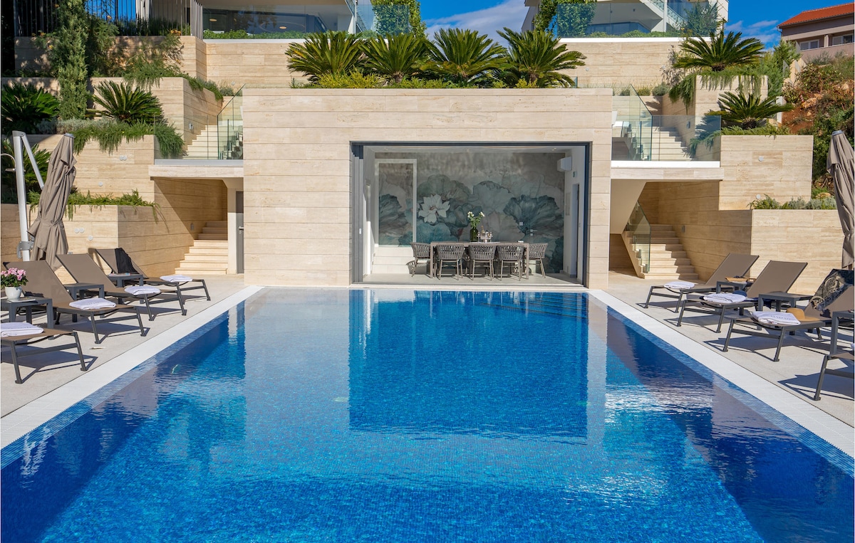 A large outdoor pool with a deep blue surface is framed by a spacious sun terrace. Sun loungers are arranged along the edge, providing a relaxing space. Lush greenery and flanking palm trees enhance the serene surroundings, while steps lead to a stylishly designed entrance area.