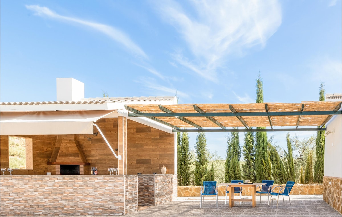 An outdoor kitchen features a rustic wood finish and an open area shaded by a pergola. Blue chairs are arranged around a wooden table, creating a comfortable dining space. Lush greenery is visible in the background, enhancing the outdoor experience in a sunny environment.