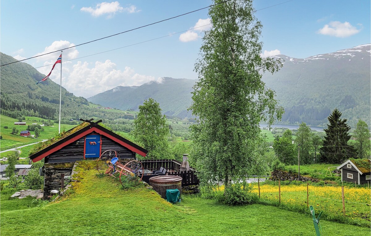 A traditional wooden house is set against a backdrop of lush green hills and distant mountains. The property features a vibrant garden with a variety of trees and flowers. A flag is seen flying, and a wooden terrace is visible, enhancing the outdoor charm.