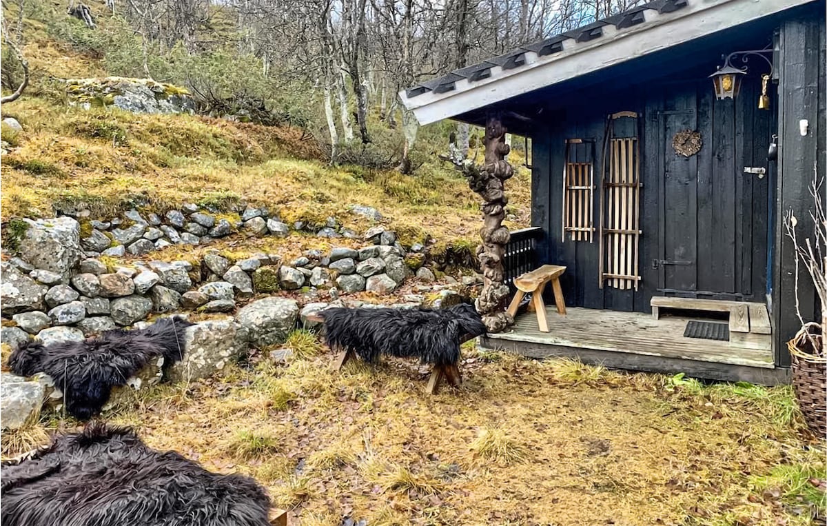 The entrance to a rustic cabin features wooden doors and a simple porch area. Black fur rugs are visible on the grassy ground, with a stone wall serving as a backdrop. Sparse trees and vegetation surround the cabin, blending naturally with the environment.