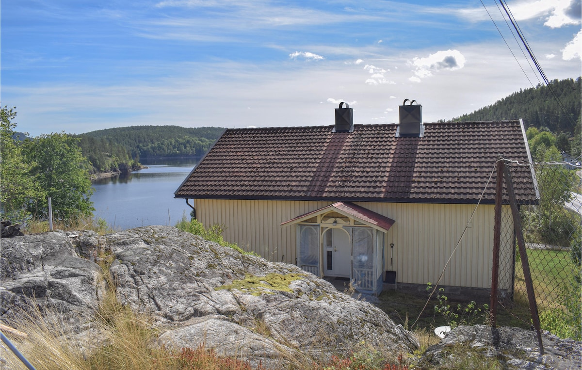 A cozy cottage, showcasing a sloped roof and light-colored exterior, is situated beside Tokke Lake. The exterior features an entrance with a small porch, framed by greenery and rocky terrain, with scenic water views visible in the background.