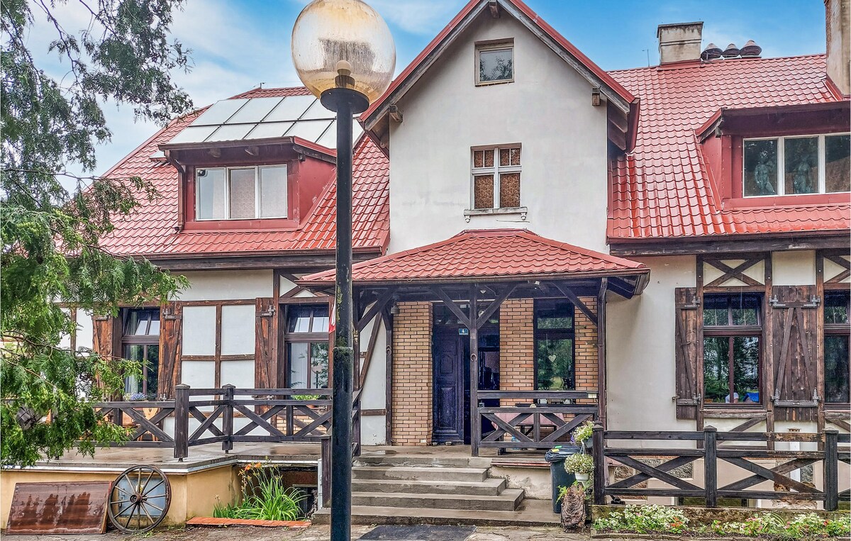 The exterior of the cottage displays a traditional design with a red-tiled roof and wooden details. A welcoming porch is framed by steps leading to the entrance, flanked by large windows. Lush greenery surrounds the property, and a light fixture stands at the forefront.