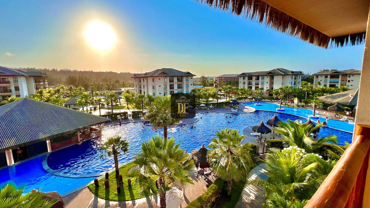 A wide view reveals the expansive pool area surrounded by lush palm trees and resort buildings. The sun sets in the background, casting a warm glow over the landscape. Leisure areas with umbrellas and lounge chairs promote relaxation by the water.