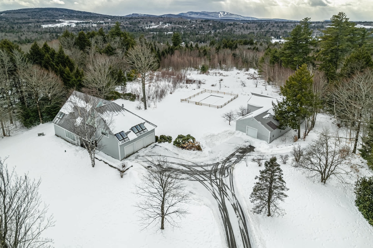 The aerial view captures a spacious property blanketed in snow, surrounded by trees and open land. A large, modern house can be seen alongside a garage, with a cleared driveway leading to them. A fenced area is also visible in the distance.