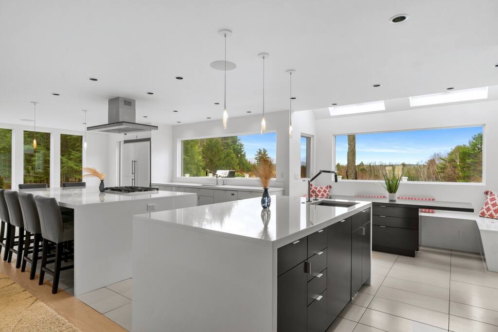The modern kitchen features sleek cabinetry and an expansive island with a white countertop. Large windows allow natural light to fill the space, offering views of the surrounding greenery. A set of black barstools is positioned at the island, enhancing the open layout.
