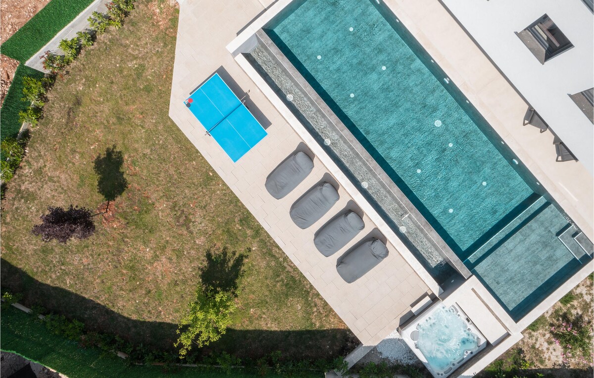 An aerial view highlights the expansive infinity pool surrounded by comfortable loungers. A sunbathing area features a vibrant blue table tennis surface, while lush greenery encircles the outdoor space, providing a serene atmosphere for relaxation.
