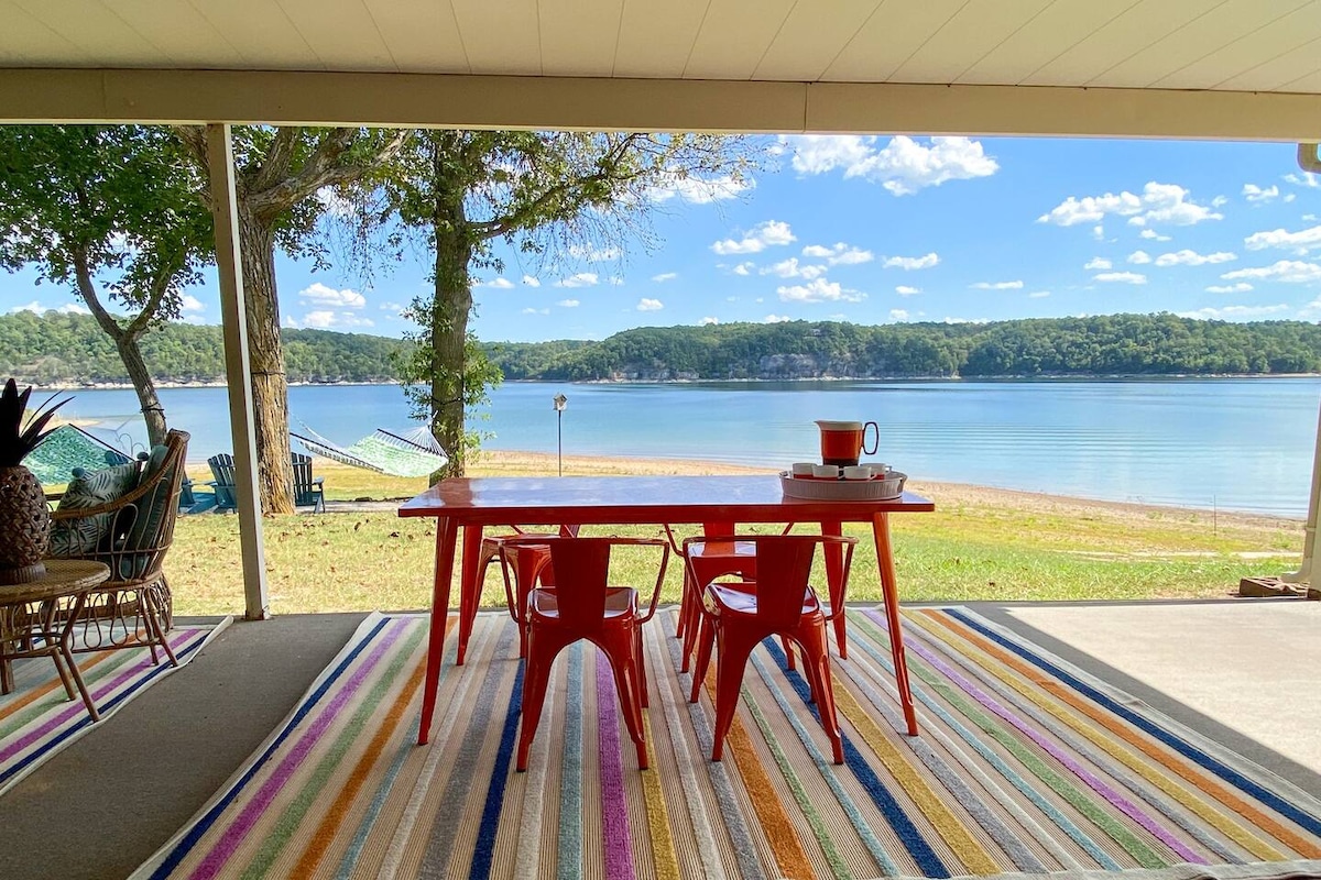 A covered patio area presents a vibrant scene with a red table and two chairs facing the waterfront. A colorful striped rug enhances the space, while the serene lake and trees are visible in the background under a clear blue sky.
