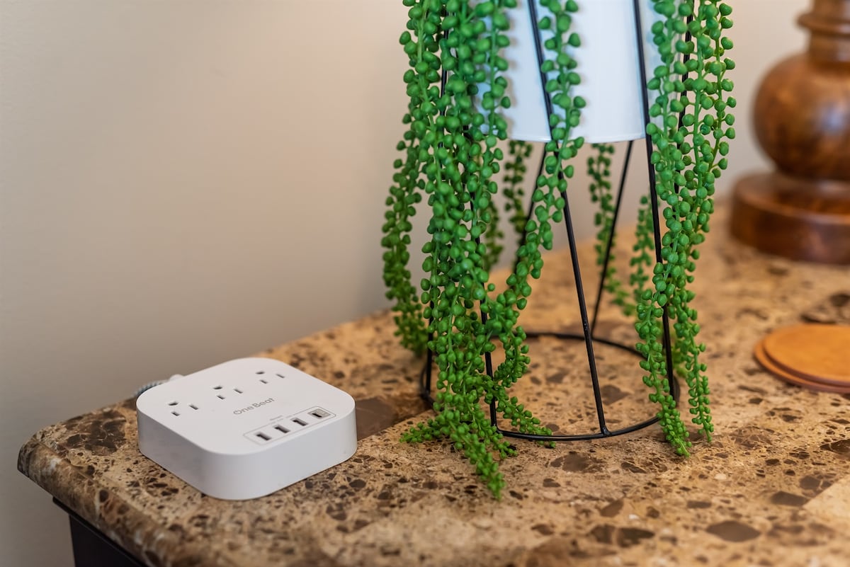A bedside table features a modern power strip with multiple outlets and USB ports. A decorative potted plant with cascading green leaves adds a natural touch, while the table's marble surface reflects light, creating a clean and functional space for charging devices.