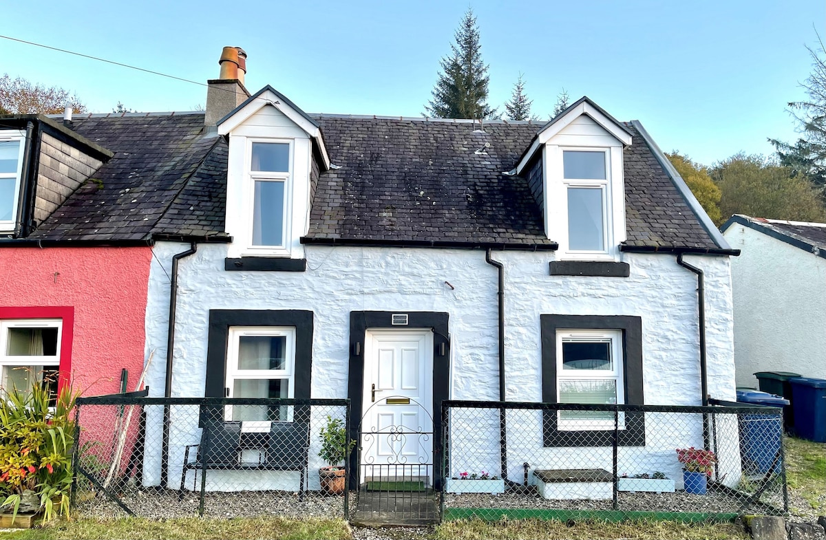 The exterior of Chapelton Cottage is displayed in a charming terrace style, featuring a white facade with black trim. The welcoming front door is adorned with a decorative design. Surrounding the entrance is a small fenced garden, with a few potted plants visible.