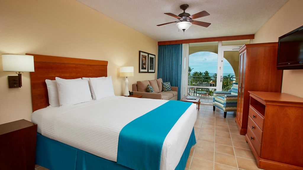 The spacious studio suite features a queen bed with crisp white linens and a turquoise accent blanket. A comfortable sofa is positioned nearby. A flat-panel TV is mounted on the wall, and natural light enters through sliding glass doors, leading to a balcony with views of the outdoors.