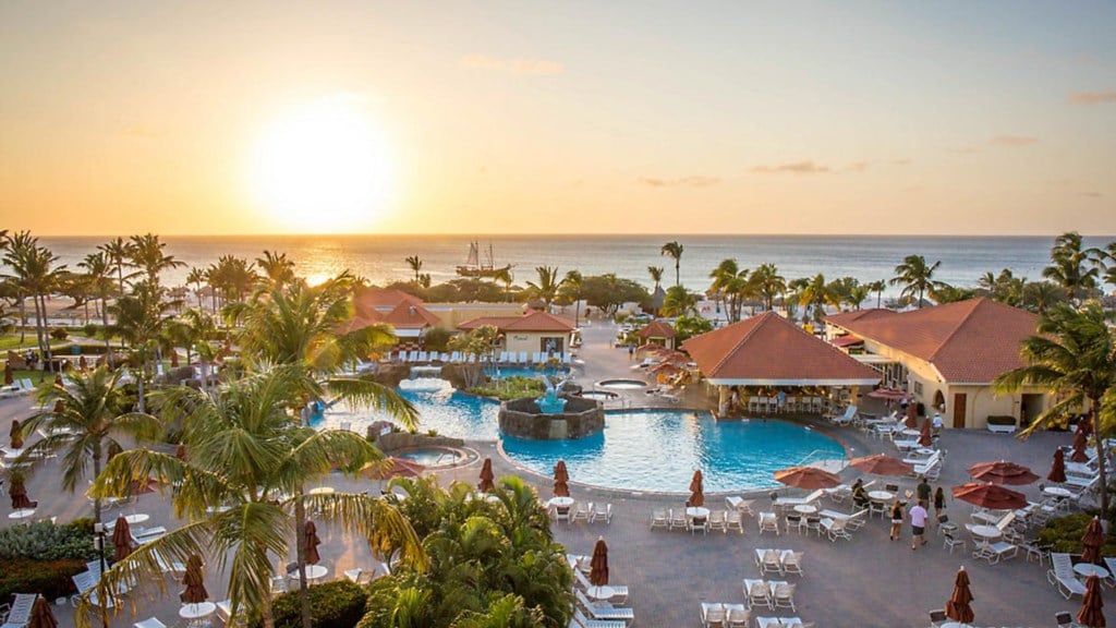 A scenic view captures the resort's expansive pool area, framed by palm trees. Lounge chairs are arranged around the pool, with umbrellas providing shade. The sun sets over the ocean, highlighting the vibrant colors of the sky and the inviting atmosphere of the resort.