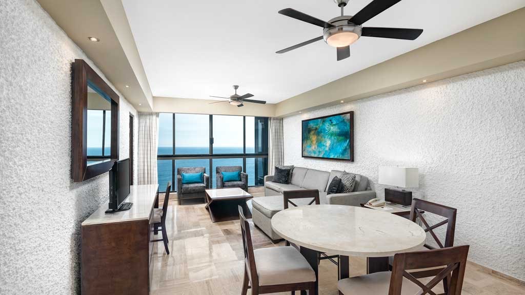 A modern living area features a comfortable sectional sofa and a round dining table surrounded by four chairs. Large windows allow views of the ocean, while a flat-screen television is mounted on the wall. Soft lighting complements the neutral color palette.