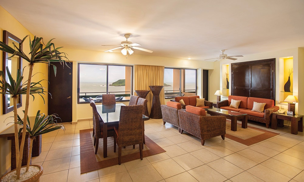 A spacious living area is filled with natural light from large windows overlooking the ocean. Comfortable seating arrangements consist of rattan furniture, including sofas and armchairs. A dining table sits nearby, complementing the inviting atmosphere of the room.