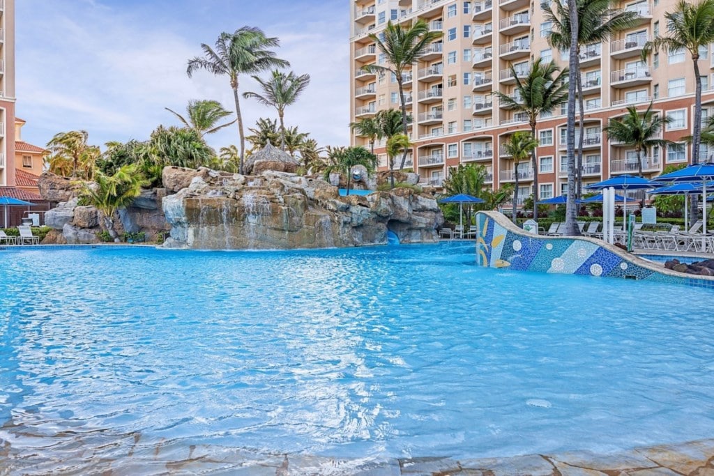 A large swimming pool is featured, surrounded by palm trees and natural rock formations. Bright blue waters reflect the sky, while a gentle waterfall cascades down rocks. Loungers with umbrellas are positioned around the pool, offering shaded relaxation areas.