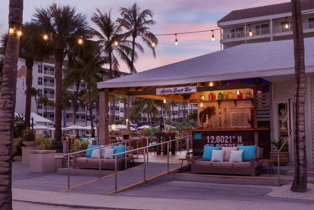 The outdoor beach bar features a wooden structure with a thatched roof, surrounded by lush palm trees. Comfortable seating with blue cushions invites relaxation. Warm ambient lighting enhances the welcoming atmosphere as the sky transitions to twilight.