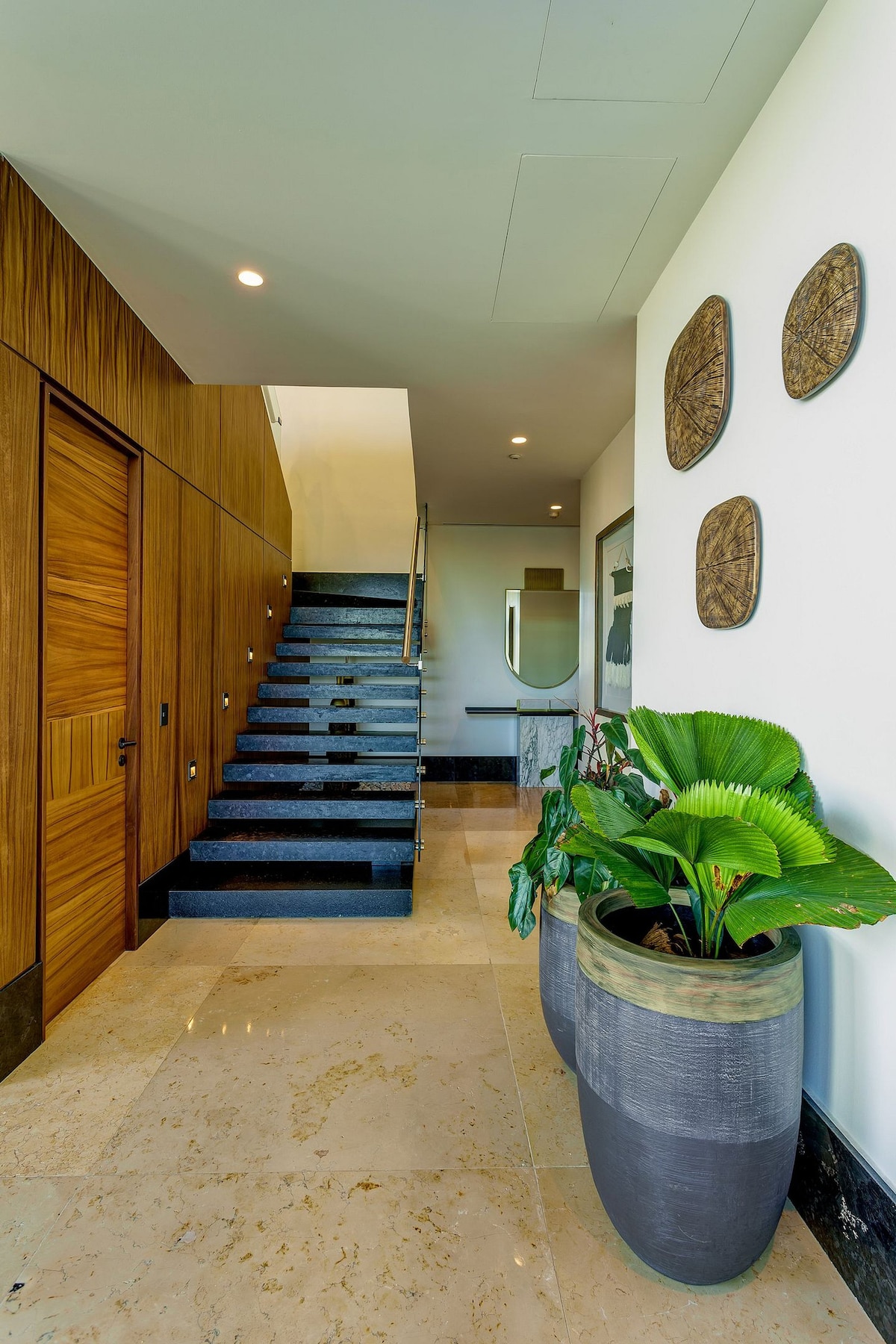 A modern entryway features a staircase with dark wooden steps and a sleek railing. The walls display decorative pieces, while a large planter holds lush green foliage. Elegant marble flooring reflects soft lighting from above.